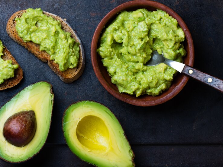 Draufsicht: Zwei Avocado-Hälften liegen auf einem dunklen Hintergrund. Daneben steht eine Guacamole-Soße in einer Tonschüssel. Auch zwei Avocado Sandwiches liegen auf dem Untergrund.