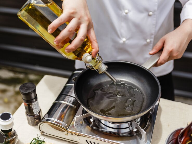 Draufsicht: Auf einer Profi-Gas-Kochplatte steht eine schwarze Pfanne, in die ein Profi-Koch Olivenöl schüttet. Neben der einzelnen Herdplatte stehen eine Pfeffermühle sowie eine weitere Flasche mit einem Gewürz.