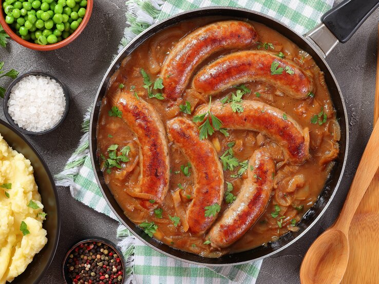 Eine Pfanne mit Bratwürsten in einer braunen Zwiebel-Biersoße. Die Pfanne steht auf einem grün-weiß karierten Geschirrtuch. Daneben ist Salz, Pfeffer, Erbsen und Kartoffelpüree.