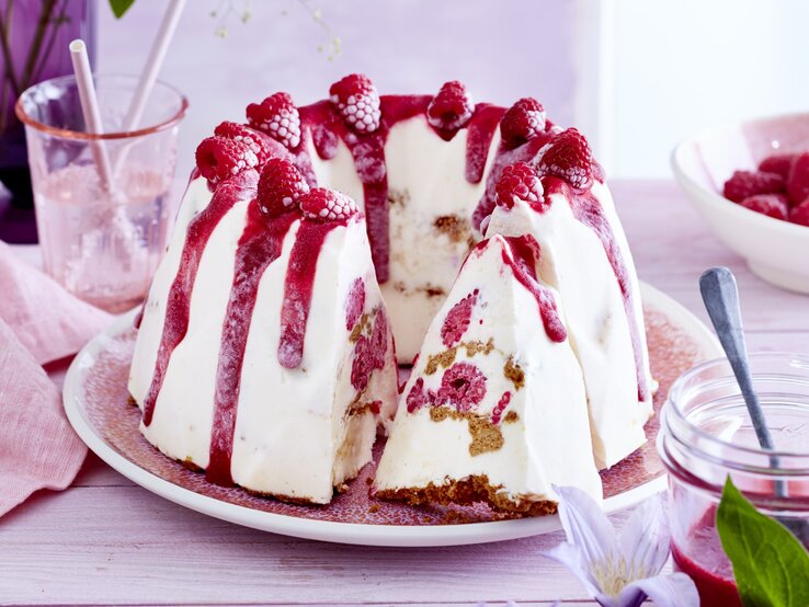Ein Traum von Dessert: Probieren Sie unseren Cheesecake-Eis-Gugelhupf! Cheesecake-Eis-Gugelhupf, mit einem Boden aus knusprigem Keks und einem Topping aus Himbeersauce und frischen Himbeeren, serviert auf einem hellen Tisch auf weißem Teller.