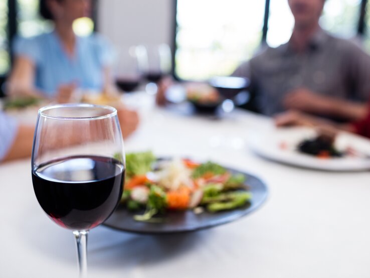 Ein Glas Rotwein im Vordergrund mit scharfem Fokus – im Hintergrund unscharf Personen beim Essen sitzen.