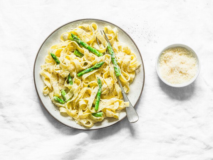 Ein Teller Frischkäse-Pasta mit grünem Spargel in der Draufsicht, daneben eine Gabel und eine Schale Parmesan.