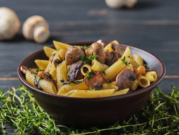 Frontalsicht: Eine dunkle Schüssel mit Penne Rigate mit Thymian-Pilzen steht auf einem dunklen Untergund aus Holz. Daneben liegen frischer Thymian und Champignons.