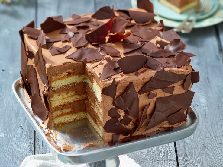 Angeschnittene viereckige Schoko-Sahne-Torte mit Schoko-Scherben verziert auf Metall-Präsentierteller, heller Hintergrund unscharf. 