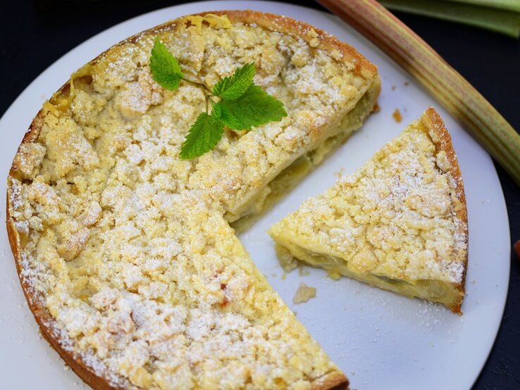 Der Rhabarberkuchen mit einem angeschnittenen Stück und ein paar Rhabarberstangen direkt von oben fotografiert. 
