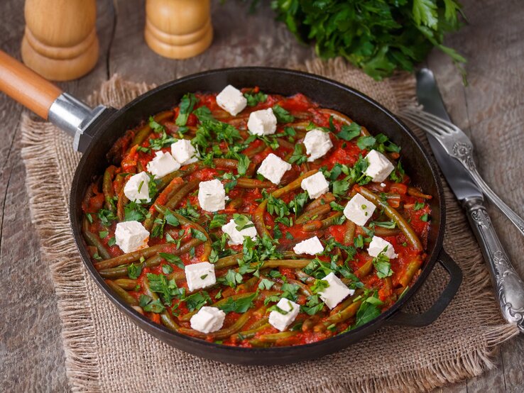 Draufsicht, schräg: eine Grüne Bohne-Pfanne mit Feta auf einem Holztisch und einem Tuch aus Leinen. Daneben liegen ein Besteck und frische Petersilie.