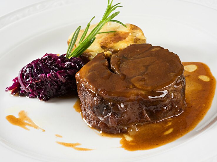 Glühweinbraten mit Soße übergossen, Rotkohl mit Rosmarinzweig und angebratener Knödel auf weißem Teller aus der Draufsicht.