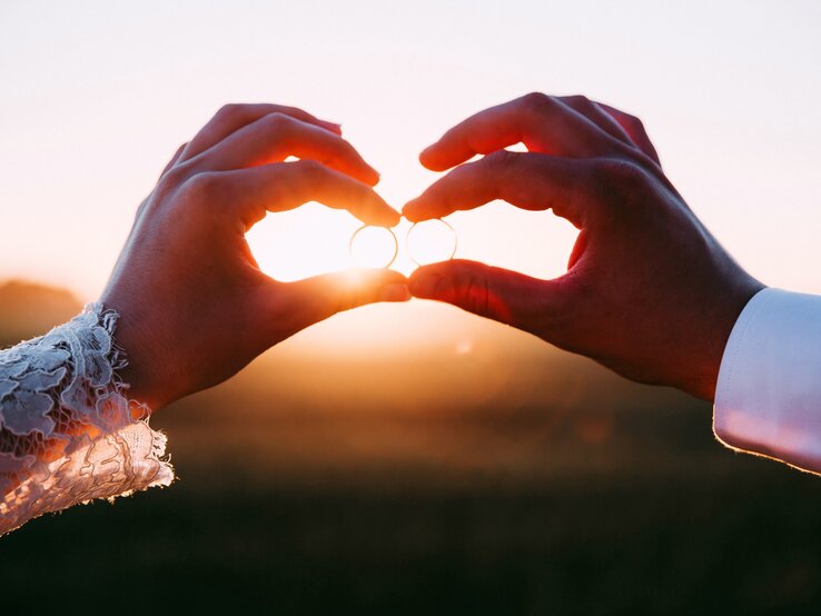 Die Hand einer Frau und die Hand eines Mannes halten Eheringe der untergehenden Sonne entgegen.