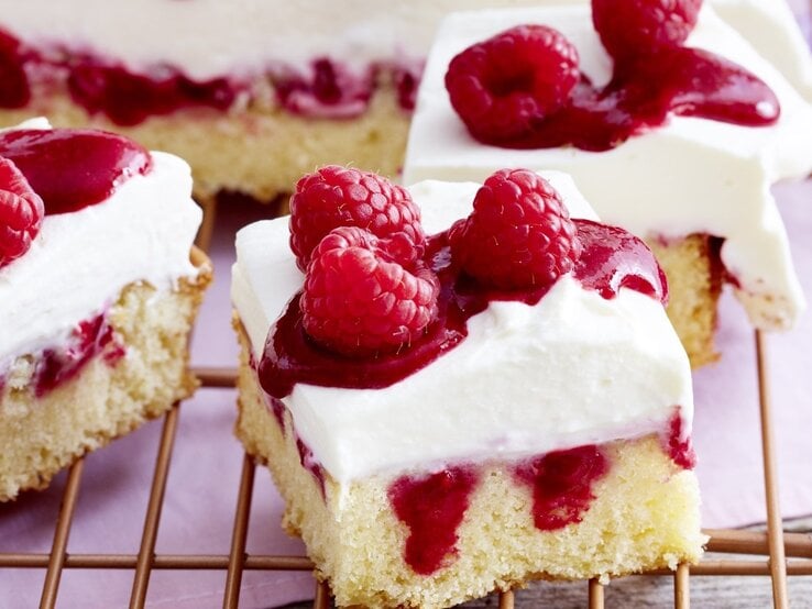 Himbeer-Mascarpone-Kuchen in eckiger Form, angeschnitten auf einem Kuchengitter auf rosanem Tischtuch.