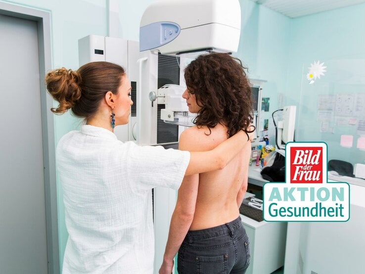 Eine medizinische Fachkraft in weißer Kleidung bereitet eine Mammographie-Untersuchung vor. Die Patientin steht ohne Oberbekleidung an dem Mammographie-Gerät und wird für die Aufnahme positioniert. Die Umgebung wirkt klinisch und professionell, mit einem hellen und sauberen Untersuchungsraum. Auf dem Bild ist auch das Logo von "Bild der Frau AKTION Gesundheit" zu sehen, was die Bedeutung von Vorsorgeuntersuchungen im Gesundheitsbewusstsein unterstreicht. Die Szene vermittelt einen wichtigen Aspekt der Frauengesundheit und präventiven Medizin.