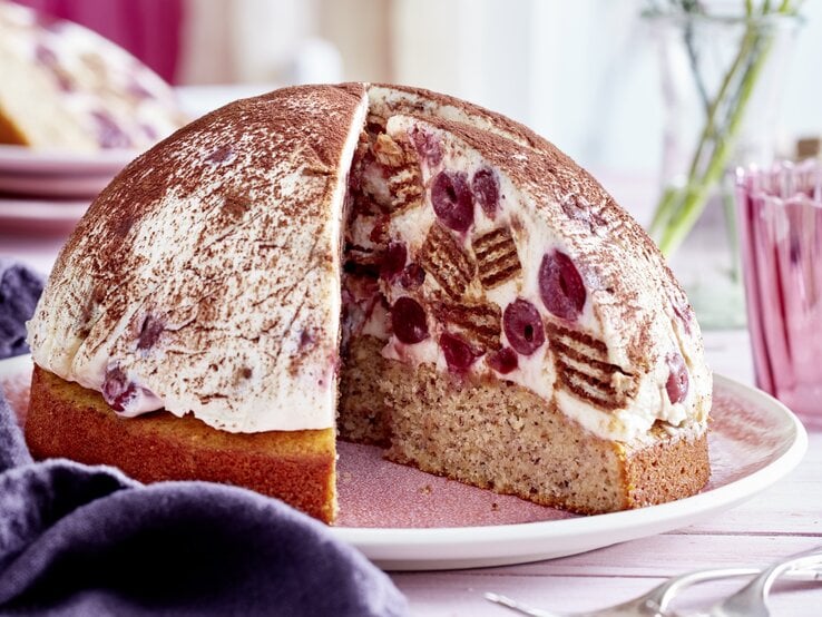 Kugelförmige Tiramisu-Kuppeltorte, gefüllt mit saftigen Kirschen und Waffeln und einem Nussboden, serviert auf einem hellen Teller vor rosarotem Hintergrund. 