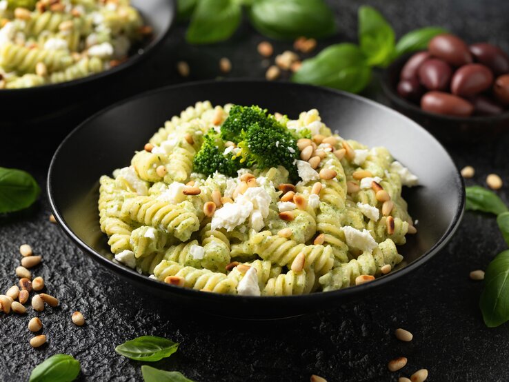 Brokkoli-Nudeln mit Feta und Pinienkernen schmecken nach Sommer pur! Brokkoli-Nudeln mit Feta und Pinienkernen auf schwarzem Teller. Dieser steht auf einer Schieferplatte. Um den Teller herum liegen frischer Basilikum und weitere Zutaten.