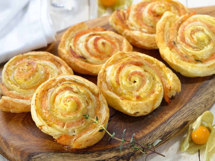 Sechs Blätterteigschnecken mit Lachs angerichtet auf einem Holzbrett. 