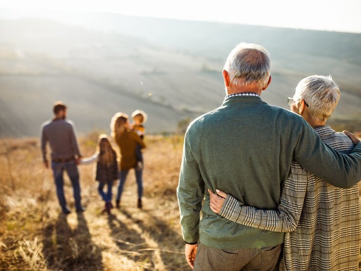 Ein älteres Ehepaar steht Arm in Arm in der Natur, die beiden schauen auf eine junge Familie vor ihnen