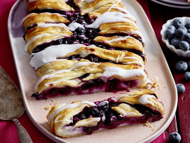 Auf einem Teller ist eine Portion Heidelbeer-Blätterteig-Striezel angerichtet. Das Gebäck ist mit einem Puderzuckerguss und Heidelbeeren überzogen. 