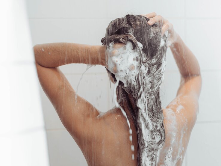Eine Person mit dem Rücken zur Kamera wäscht sich das Haar unter der Dusche. Ihr Haar ist voller Schaum, und sie hält es mit beiden Händen hoch, während Wasser daran herabfließt. Die Szene vermittelt eine Routine der Körperpflege und die ruhige Atmosphäre eines Badezimmers. Das Bild konzentriert sich auf die Aktion der Haarwäsche, wobei die genaue Uhrzeit für diese Tätigkeit nicht angegeben ist.