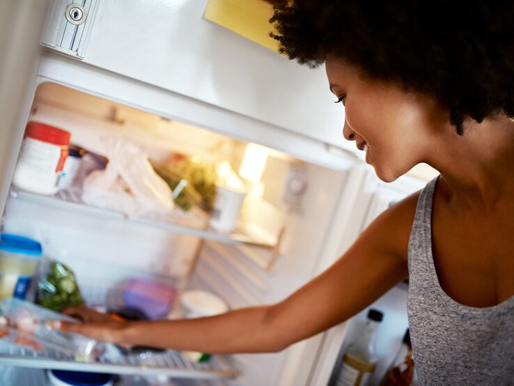 Eine Frau mit lockigem Haar öffnet einen Kühlschrank und schaut hinein. Sie steht im Vordergrund und ist seitlich abgebildet, mit einem Arm im Kühlschrank. Im Inneren des Kühlschranks sind verschiedene Lebensmittel zu erkennen, wie Gemüse, Behälter und Flaschen. Die Szene spielt sich in einer hellen Küche ab.