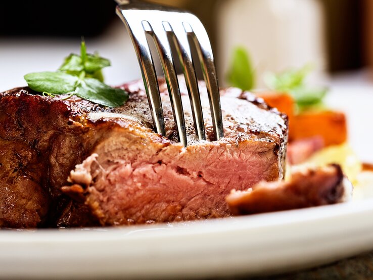 Es geht doch nichts über ein himmlisch saftiges Stück Fleisch! Und es gibt eine Zutat, die hilft dabei. Gabel steckt in einem saftigen Stück Fleisch auf weißem Teller.