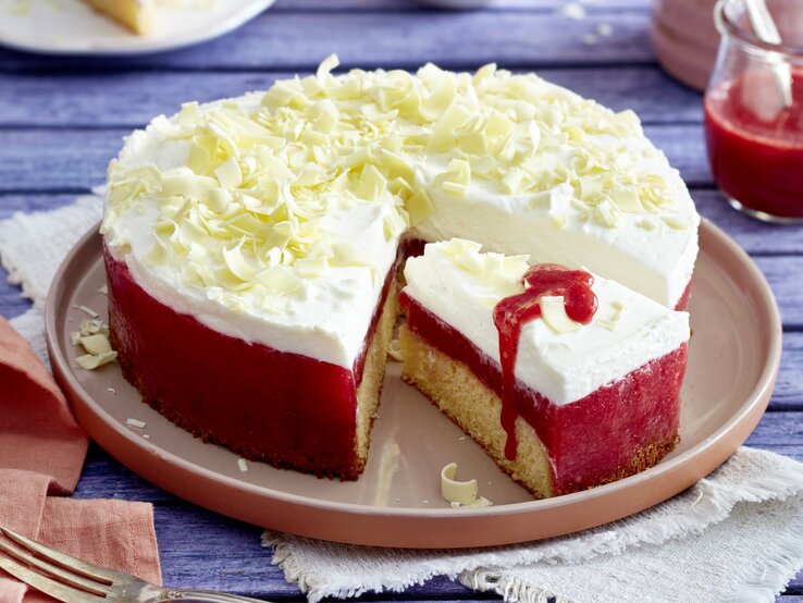 Frau-Holle-Kuchen mit Schneeflocken aus weißer Schokolade und Erdbeer-Glasur, angeschnitten serviert auf einem Kuchentablett auf blauem Holztisch. 