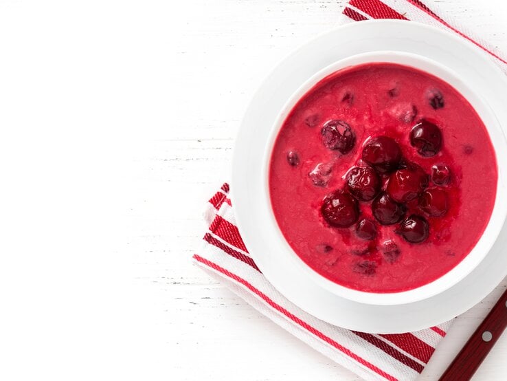 Ein weißer Teller mit der roten Suppe auf rot-weißem Tuch und weißem Tisch. Alles direkt von oben fotografiert.