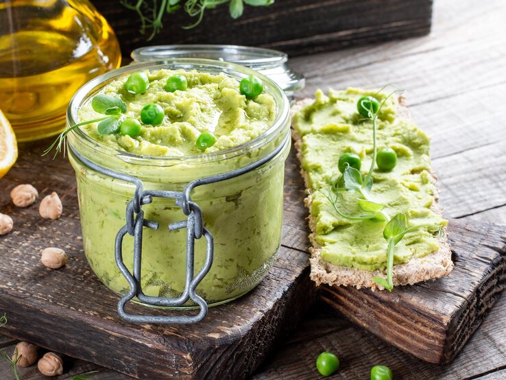 Ein Weckglas mit dem Erbsen-Hummus auf einem Holzbrett mit Zitrone und Olivenöl.