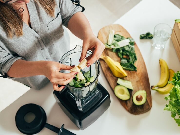 Eine Frau bereitet einen grünen Smoothie vor. Sie gibt frische Zutaten in einen Mixer, darunter Spinatblätter und Bananenstücke. Auf dem Holzbrett neben ihr liegen weitere frische Zutaten wie Avocadohälften, Bananen und Salatblätter. Die Szene spielt in einer hellen, modernen Küche. Der Fokus liegt auf den Händen der Frau und den gesunden Lebensmitteln, was eine Atmosphäre des bewussten Essens und der Gesundheitspflege suggeriert.