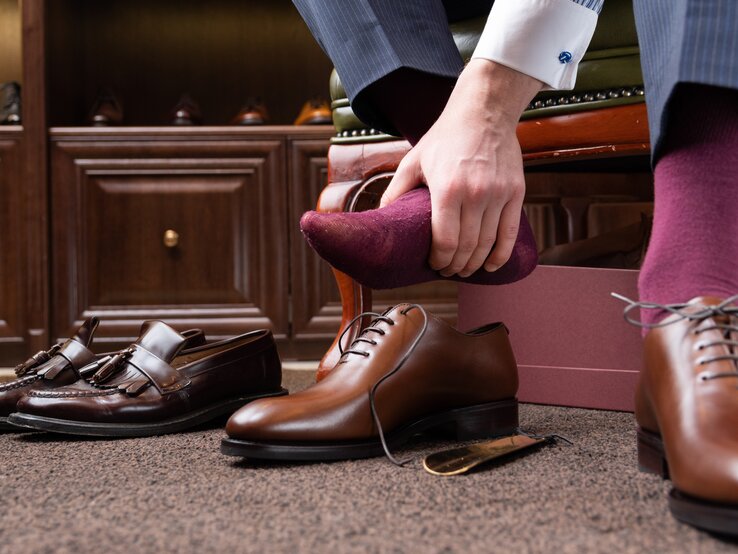Ein Mann sitzt und zieht einen burgunderroten Socken an seinem Fuß aus, um ihn offenbar gegen ein neues Paar brauner Lederschuhe zu tauschen, die neben ihm stehen. Im Hintergrund sind ein Schuhkarton und weitere Paare von hochwertigen Herrenschuhen auf einem Holzregal zu sehen.