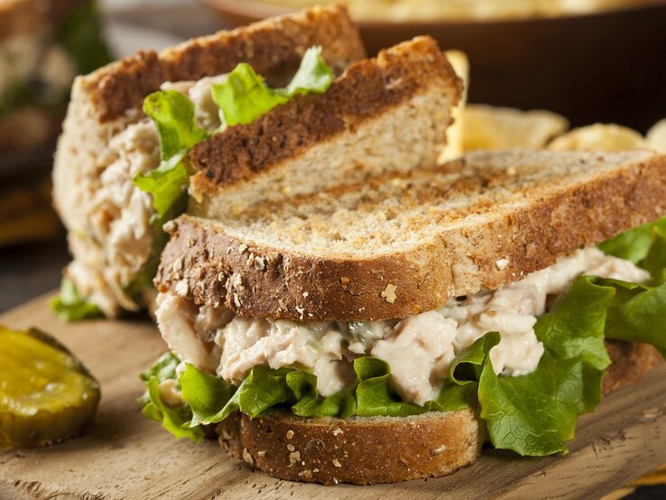Zwei Thunfisch-Sandwiches auf einem Holzbrett mit einer Gewürzgurkenscheibe und unscharfem weiteren Essen im Hintergrund.