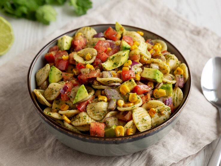 Nudelsalat-Mais-Avocado Eine Schüssel Nudelsalat mit Mais und Avocado, außerdem Paprika, drumherum frische Zutaten wie Kräuter und eine halbe Limette, außerdem 2 Löffel.