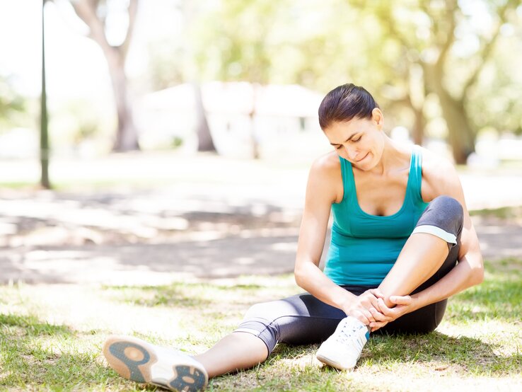 Eine Frau in Sportkleidung sitzt auf dem Gras in einem Park und berührt ihren Fußknöchel, als ob sie Schmerzen hätte. Sie trägt ein türkisfarbenes Tanktop, schwarze Sportleggings und Laufschuhe. Die Szene wirkt sonnig und friedlich, mit Bäumen und einer verschwommenen Umgebung im Hintergrund, was darauf hindeutet, dass sie sich inmitten einer entspannenden Naturumgebung befindet.