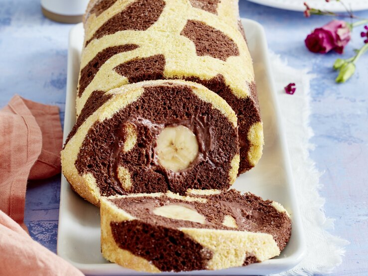 Giraffen-Bananenrolle Angeschnittene Giraffen-Bananenrolle mit Schoko-Bananen-Füllung und dunklen Punkten auf hellem Teig. Untergrund hellblau.