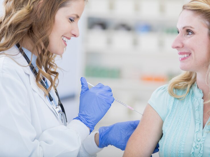 Eine Ärztin mit bräunlichen Haaren in weißem Kittel, die blaue Handschuhen trägt, gibt einer blonden Frau, die ein hellblaues Oberteil trägt eine Grippeschutzimpfung.