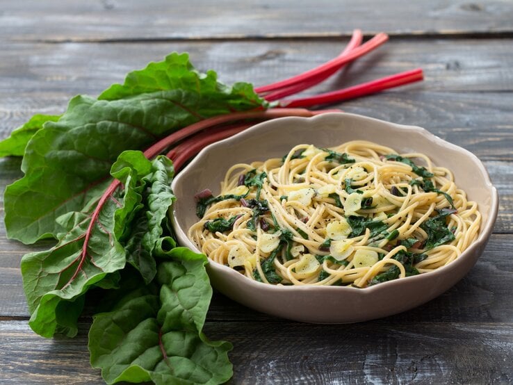 Pasta mit Mangold in cremiger Frischkäse-Soße in beigem Teller auf Holztisch. Daneben bunter Mangold. Draufsicht.