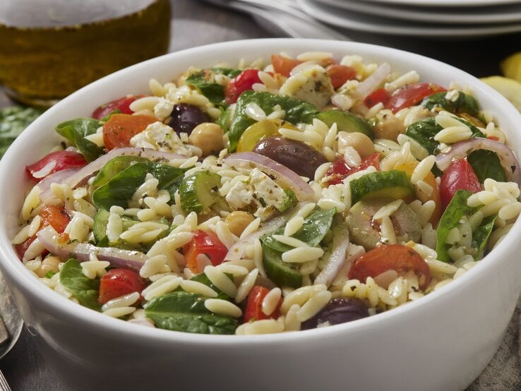 Mediterraner Orzo-Salat mit Spinat, Tomaten und Oliven in weißer Schüssel. 