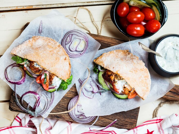 Pita-Gyros mit Beilagen auf Küchenkrepp auf hellem Untergrund. Daneben Schale mit Tzaziki und kleine Tomaten. Draufsicht.