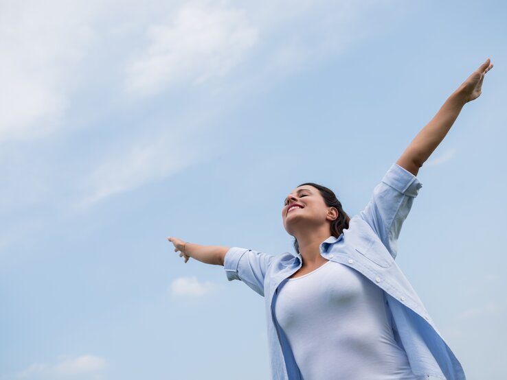  Das Bild zeigt eine Frau, die mit ausgebreiteten Armen nach oben steht und den Himmel anschaut. Sie scheint ein Gefühl von Freiheit oder Erleichterung zu erleben, vielleicht genießt sie einen Moment der Ruhe oder des Triumphes. Die Frau trägt eine blaue Bluse und ein weißes Top, und der Hintergrund besteht aus einem klaren blauen Himmel mit einigen leichten Wolken. Die Pose der Frau und ihr Gesichtsausdruck vermitteln ein starkes Gefühl der Freude und der Offenheit gegenüber der Welt um sie herum.