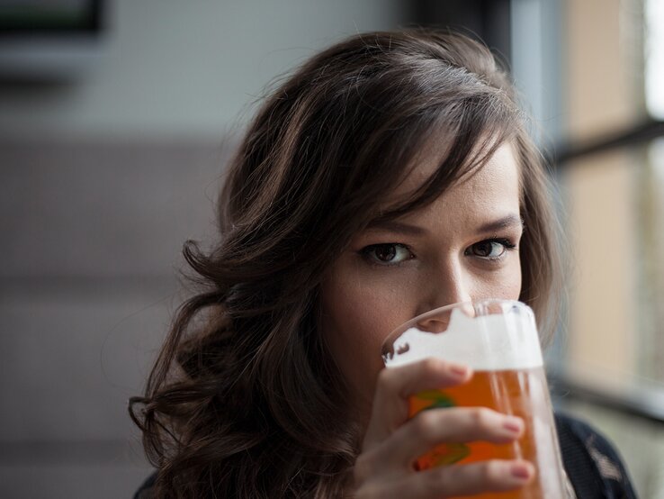Eine Frau mit lockigem braunem Haar hält ein Glas Bier & trinkt, während sie direkt in die Kamera blickt.