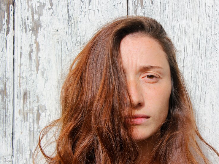 Porträt einer Frau mittleren Alters mit ernstem Gesichtsausdruck vor einer abblätternden weißen Holzwand. Ihre braunen Haare fallen locker über die rechte Seite ihres Gesichts, wodurch eines ihrer Augen verdeckt wird. Auf ihrer Stirn zeichnen sich leichte Zornesfalten ab. Das Bild vermittelt eine Atmosphäre von Nachdenklichkeit oder Sorge.