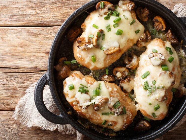 Überbackene Hähnchenbrust mit Pilzen, Lauch und Mozzarella in schwarzer Gussform mit Henkeln auf Leinentuch und Holztisch. Aufnahme aus der Vogelperspektive.  