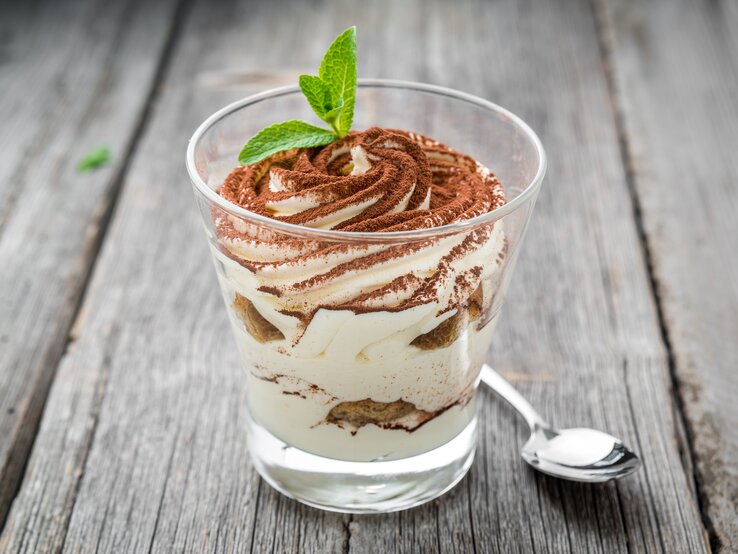 Milchmädchen-Tiramisu Ein durchsichtiges Glas gefüllt mit einem Dessert bestehend aus gezuckerter Kondensmilch (Milchmädchen) und Gebäck. Das Ganze steht auf einem grauen Holztisch.