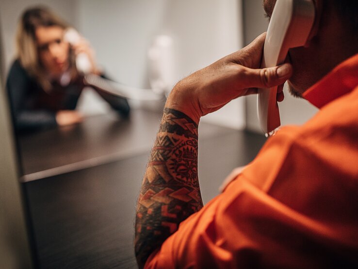 Ein Häftling in orangefarbenem Anzug telefoniert im Knast durch eine Glasscheibe getrennt mit einer Besucherin