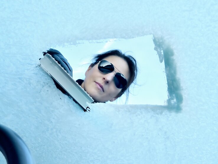 Blick aus dem Inneren durch die Frontscheibe eines Autor, während eine Frau den Schnee entfernt. 