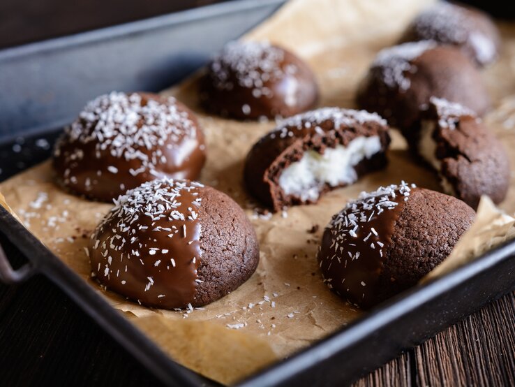 Schokocookies mit Kokosfüllung, Schokoglasur und Kokosflocken auf einem Backblech mit Backpapier auf einem dunklen Holztisch. 