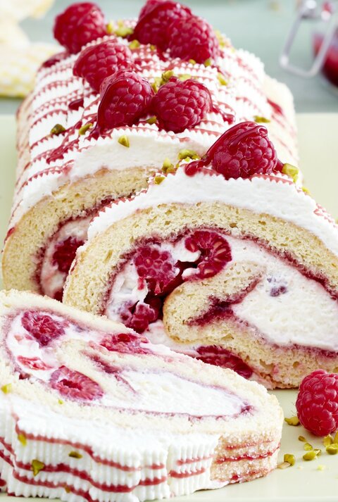 Eine sahnige Himbeerrolle mit Schmand liegt auf einer rechteckigen Porzellanplatte. Drumherum liegen Himbeeren und Servietten.