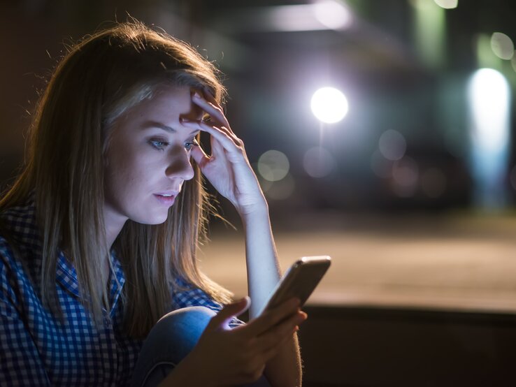 Junge Frau schaut in einem dunklen Zimmer überrascht auf ihr Handy