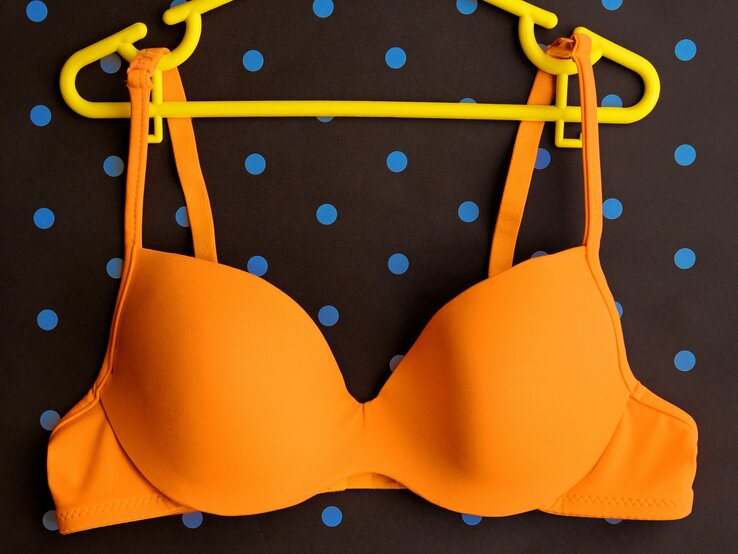 Leuchtend orangefarbenen BH, der an einem gelben Kleiderbügel hängt. Der Hintergrund ist dunkel mit blauen Punkten.