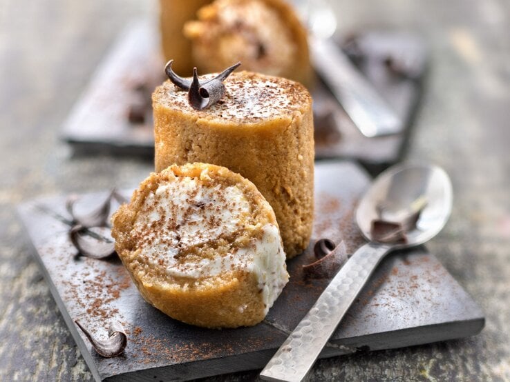 Tiramisu-Röllchen auf einem quadratischen schwarzen Brett. Daneben liegt ein Löffel. 