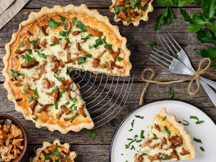 Mehr Aroma geht nicht: Eine herrlich duftende Pfifferling-Quiche mit Schmand und Bergkäse. Eine angeschnittene Pfifferling-Quiche mit Schmand und Bergkäse steht auf einem Kuchengitter. Daneben liegen Besteck sowie ein kleiner Teller mit einem Stück Quiche.