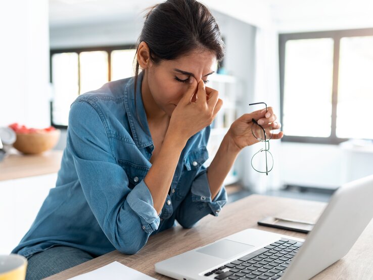 Eine Frau in einem blauen Jeanshemd sitzt vor einem Laptop und hält ihre Brille in einer Hand, während sie mit der anderen Hand ihre Nasenwurzel massiert, ein Zeichen von Stress oder Ermüdung. Vor ihr auf dem Tisch liegen ein Notizblock und ein Smartphone. Der Raum ist hell und modern eingerichtet, mit großen Fenstern, die für natürliches Licht sorgen. Die Atmosphäre scheint ruhig, doch die Frau wirkt nachdenklich oder gestresst.