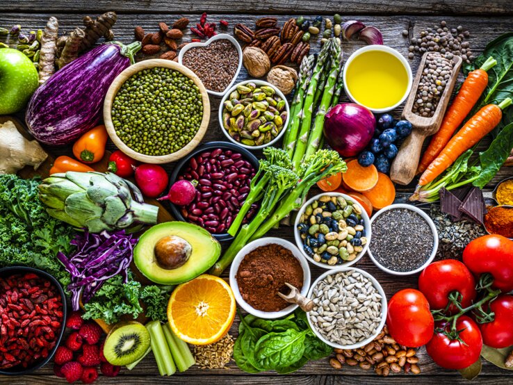 Ein reichhaltiges und farbenfrohes Arrangement von Superfoods und gesunden Lebensmitteln auf einem rustikalen Holztisch. Zu den sichtbaren Lebensmitteln gehören verschiedene frische Gemüsesorten wie Auberginen, Tomaten und grünes Blattgemüse, Obst wie Äpfel und Beeren, Nüsse, Samen, Hülsenfrüchte und Gewürze. Das Bild strahlt Frische und Vielfalt aus und betont eine nährstoffreiche Ernährung.